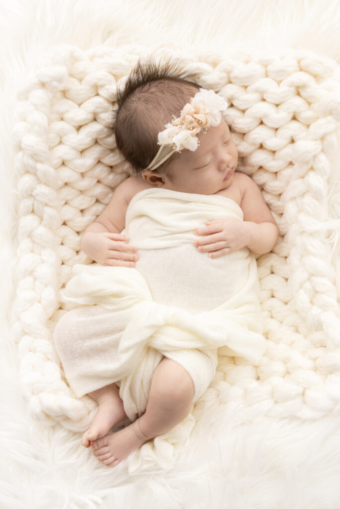 in this vertical newborn portrait, Baby Chloe sleeps peacefully with her hands resting in a relaxed way across her torso; she is lightly wrapped in ivory and wears a stretchy floral headband