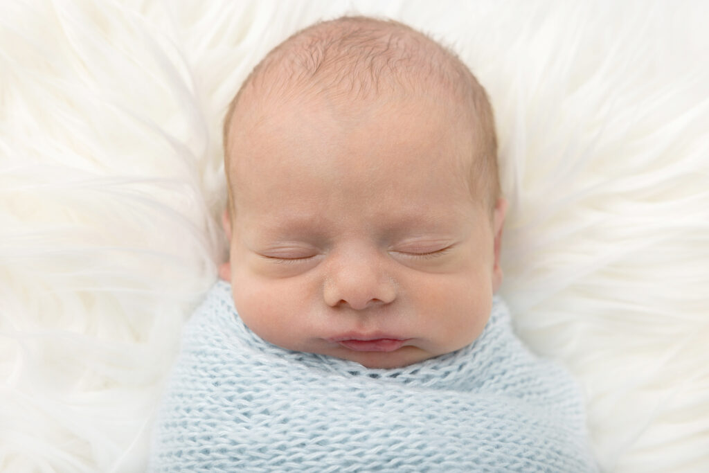 A newborn twin boy with peach fuzz hair sleeps with his sweetly drooping mouth and chin resting on a light blue textured blanket, as he lies on a white flokati.