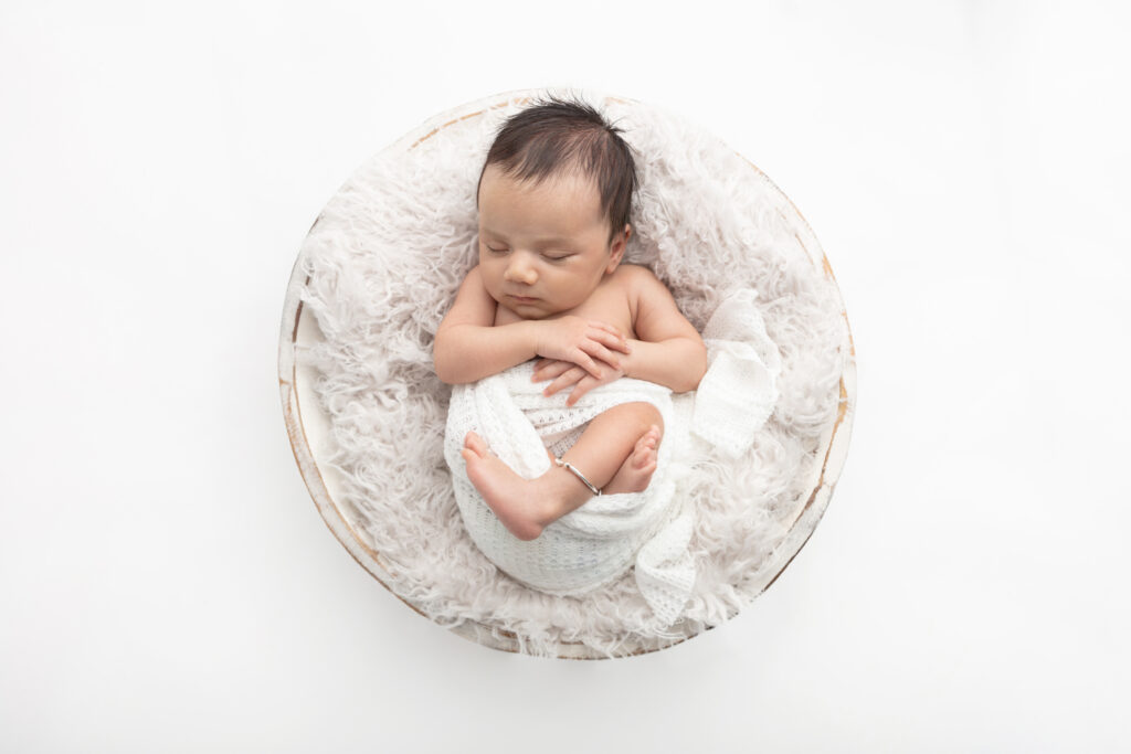 A newborn baby boy with long, delicate arms, legs, fingers, and toes, lies in a relaxed sleeping pose in a whitewashed wash basin. He wears a traditional Indian black beaded bangle around his ankle.
