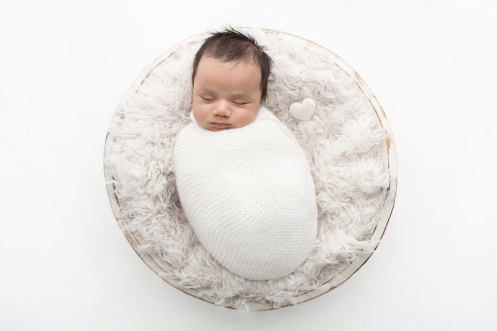 A newborn baby boy is tightly swaddled in a textured white blanket. He lies on a white flokati in a rustic whitewashed wash basin, with a felted white heart laying next to him.