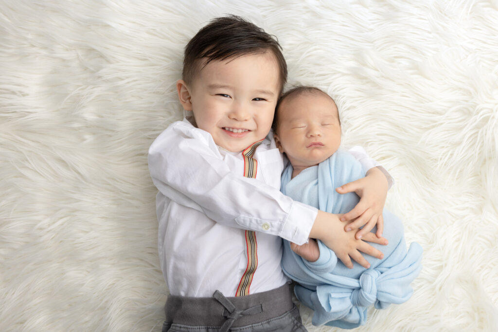 a newborn baby boy and his big brother are photographed by Karen Kahn, of Looking Up Photography; the pair are lying on an ivory colored flokati