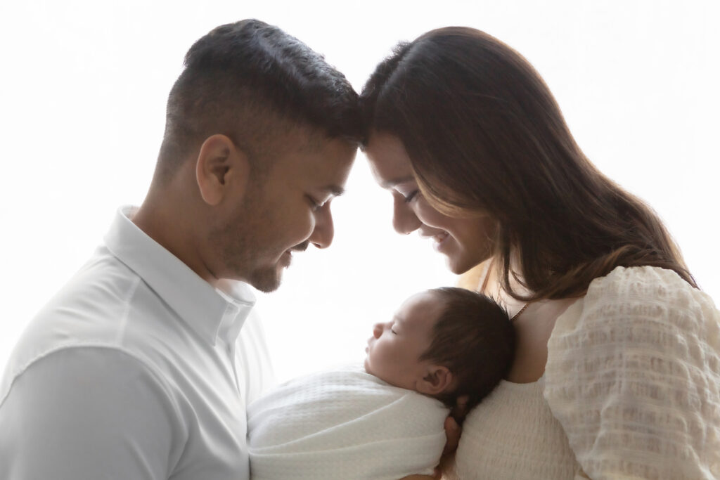 A new father and mother are photographed, backlit by the natural light of the Looking Up Photography studio. They hold their newborn son between them and look down at him, smiling.
