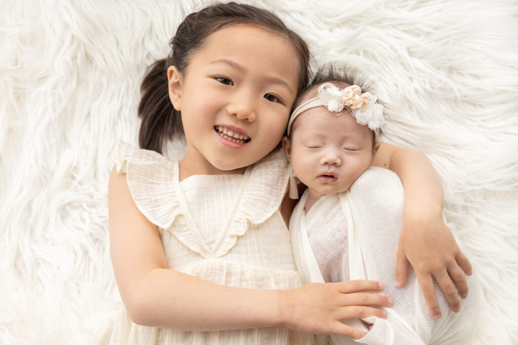 five year-old big sister Emma happily lies on an ivory flokati with her arm around her newborn baby sister, Chloe. the girls wear shades of white and ivory