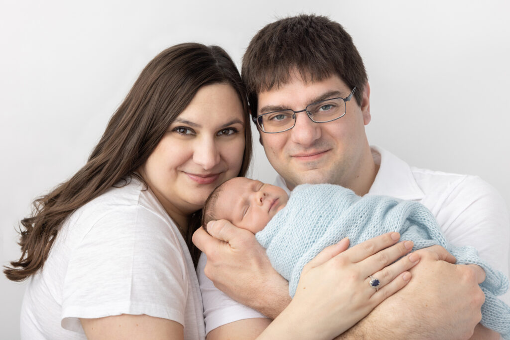 New twin parents Leah and Patrick hold one of their newborn twin boys and smile gently at the camera. The baby boy is wrapped in light blue and sleeps peacefully.