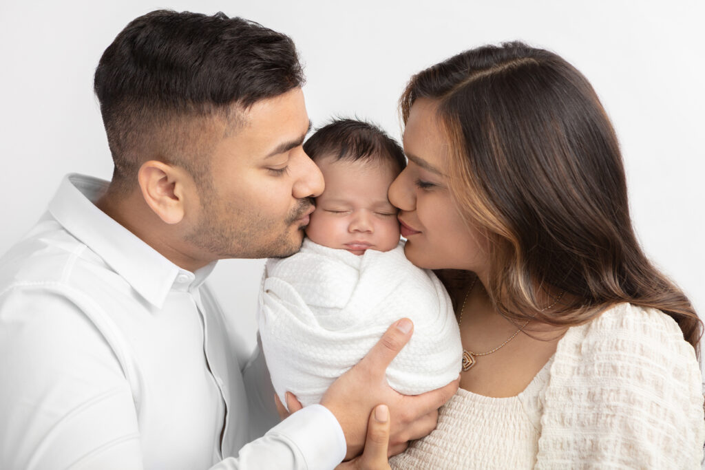 A young Indian couple hold their newborn son between them and kiss his cheeks as he sleeps, swaddled tightly in a textured white blanket. The trio are photographed at the Looking Up Photography studio in Greenwich, Connecticut.