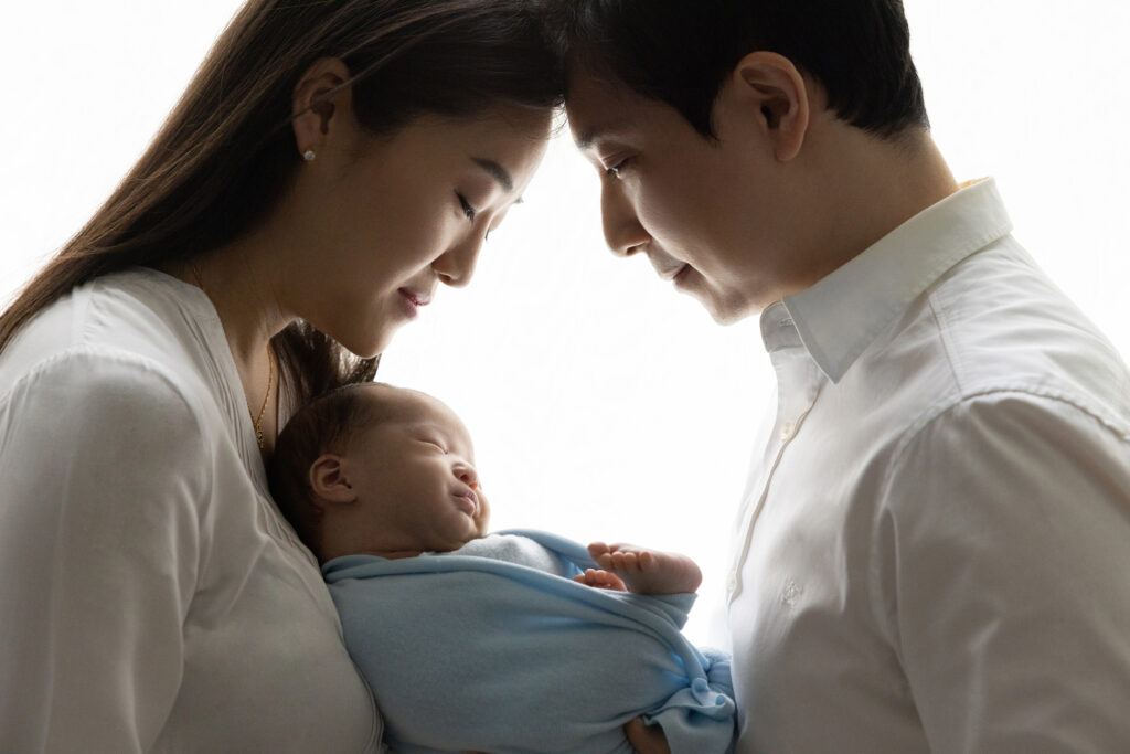 new parents are photographed backlit, semi-silhouetted, holding their newborn baby boy in between them; the profiles of mom, dad, and baby are visible