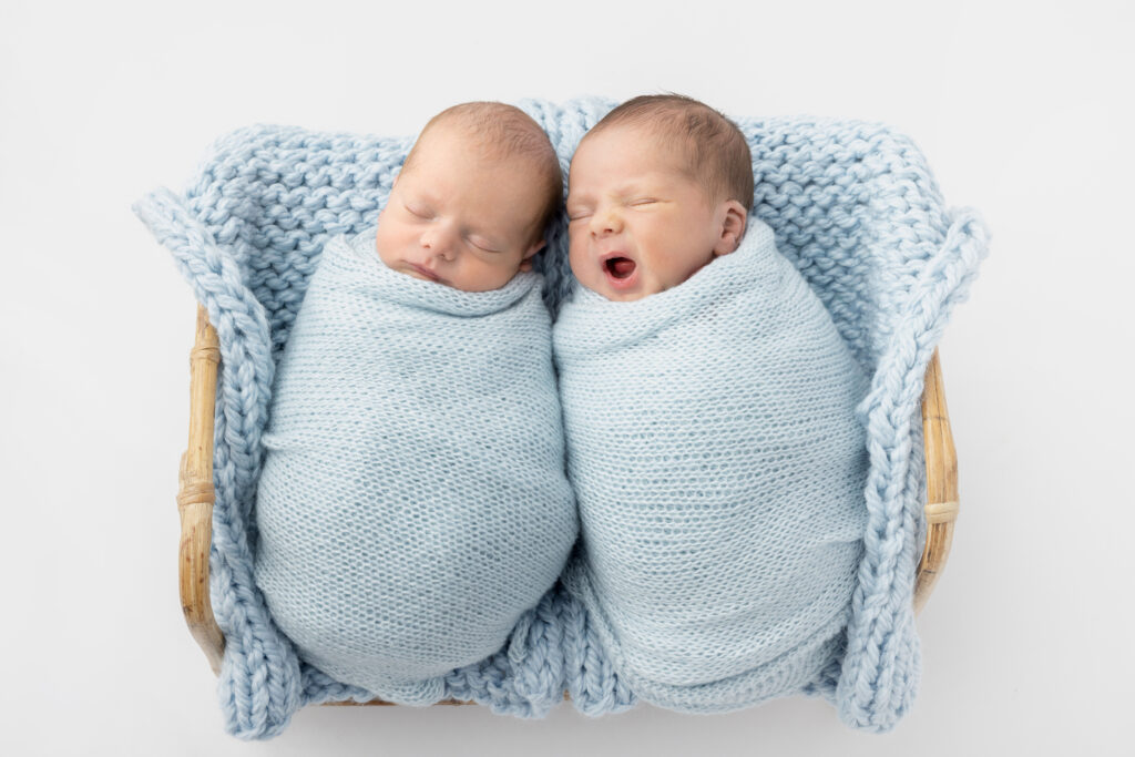 Newborn twin boys, tightly wrapped in light blue, are photographed on a textured, light blue blanket square in a light colored bamboo basket. One of the baby boys is yawning.