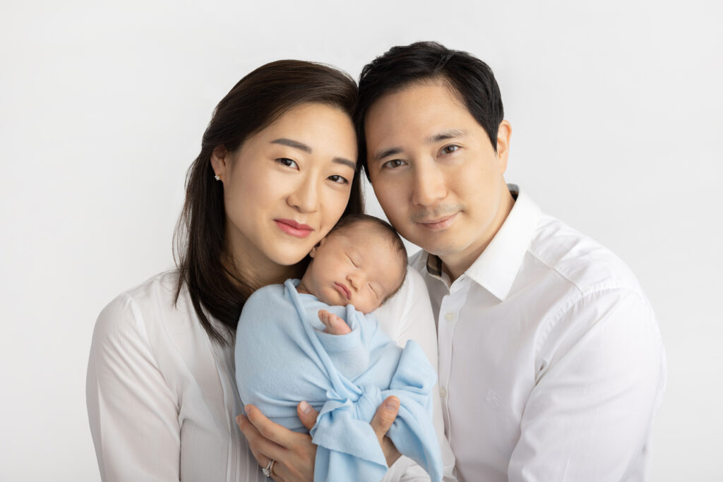 new parents are photographed in the Looking Up Photography studio in Cos Cob, Greenwich, Connecticut, by Karen Kahn; the duo wear white shirts and hold their son in between them