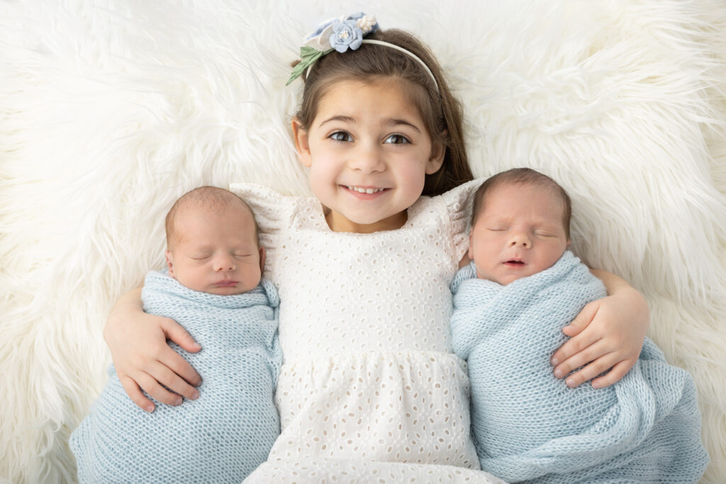 Four year-old big sister Elodie wears an eyelet dress and smiles with enthusiasm as she lies on a creamy colored flokati and holds her newborn twin brothers in either arm.