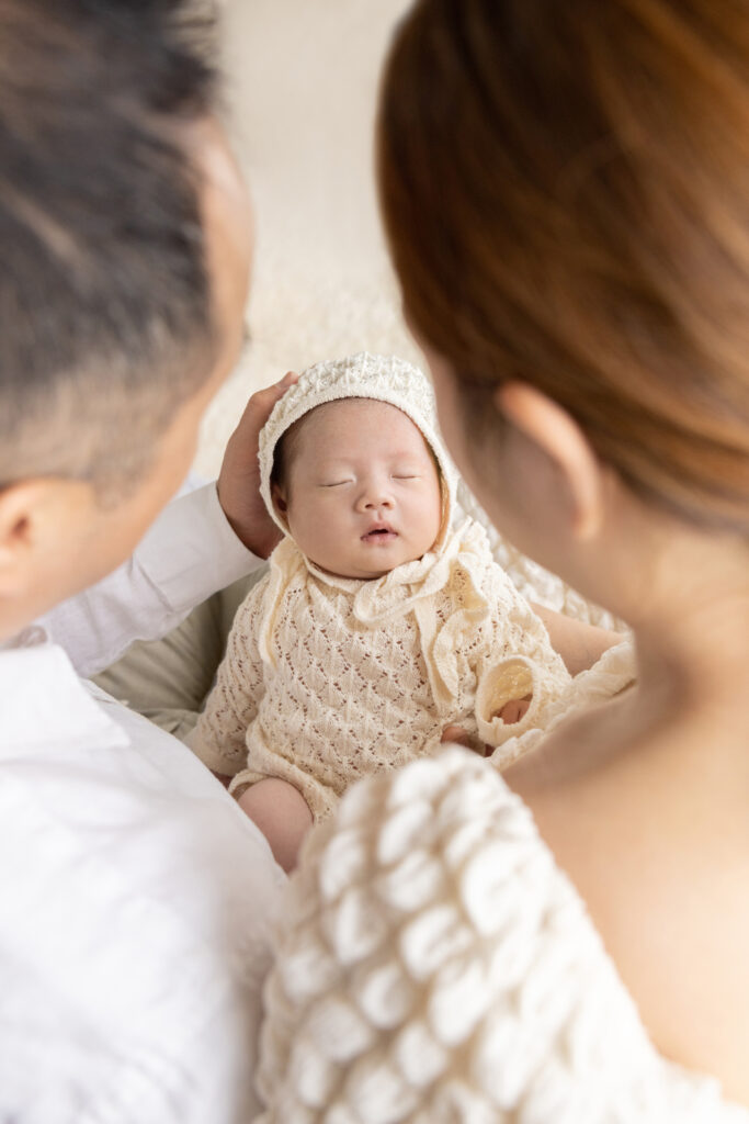 in this newborn photo from a unique perspective, the photographer's lens peers through the back of the heads of mom and dad, who hold their newborn baby girl in their hands and laps
