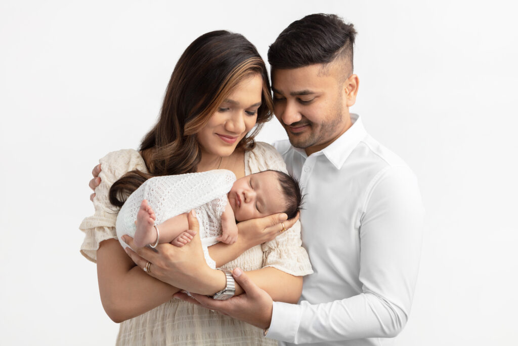 A lovely young Indian couple hold their sleeping newborn son in their arms and look down at him, smiling, as the baby boy relaxes in his mother's arms. The trio are photographed by Karen Kahn, of Looking Up Photography.