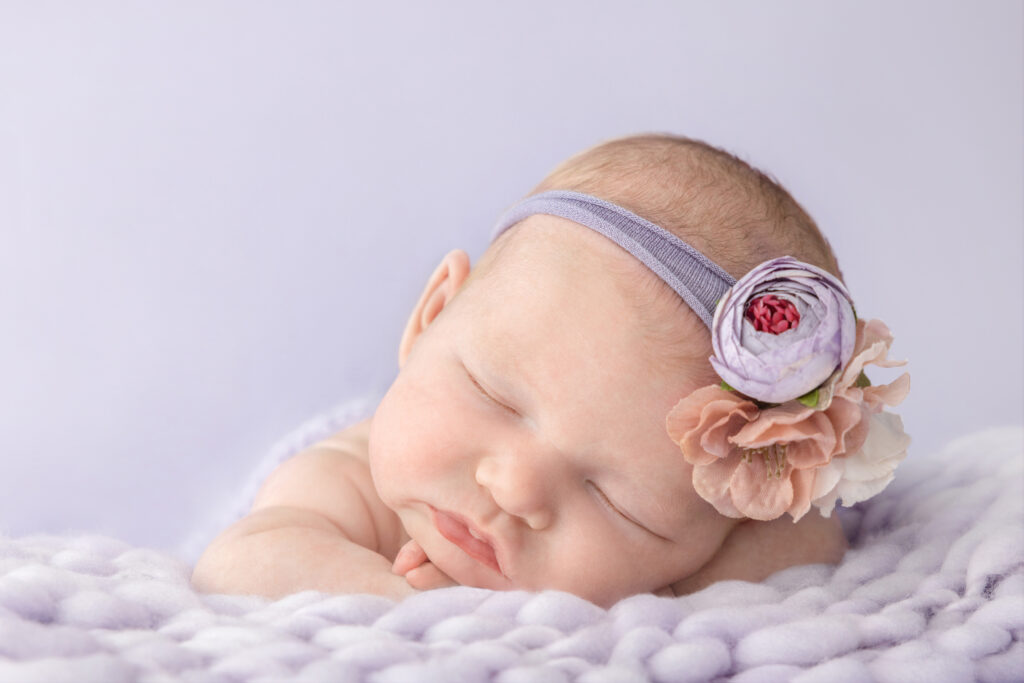 A newborn baby girl sleeps peacefully, her arms and hands folded underneath her as she lies on a plush, oversized knit blanket in pale purple. The baby wears a faux floral stretchy headband.