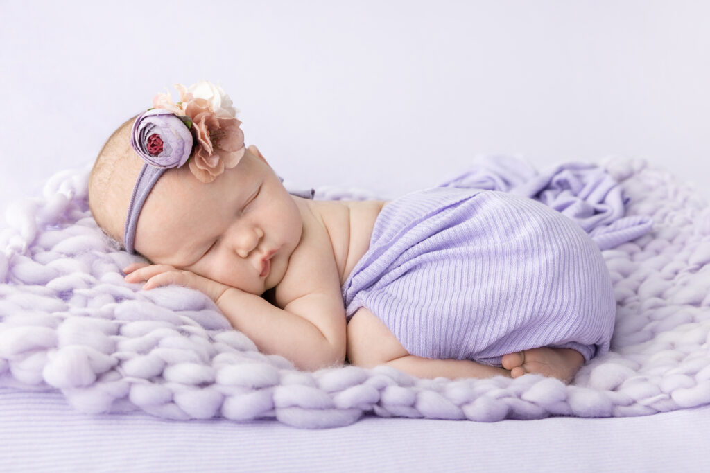 A newborn baby girl lies partially on her side on a plush knit blanket, which is on another, larger, ribbed blanket in a pale purple. She wears a stretchy floral headband of pale purple, blush, and white flowers.
