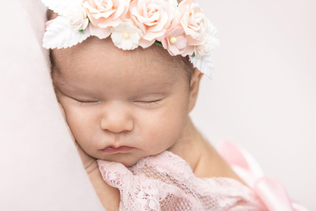 A close up photo of a newborn baby girl named Fallon. Fallon wears a floral crown of white and peachy pink beautifully crafted faux flowers, and lace wrapped delicately around her shoulder.