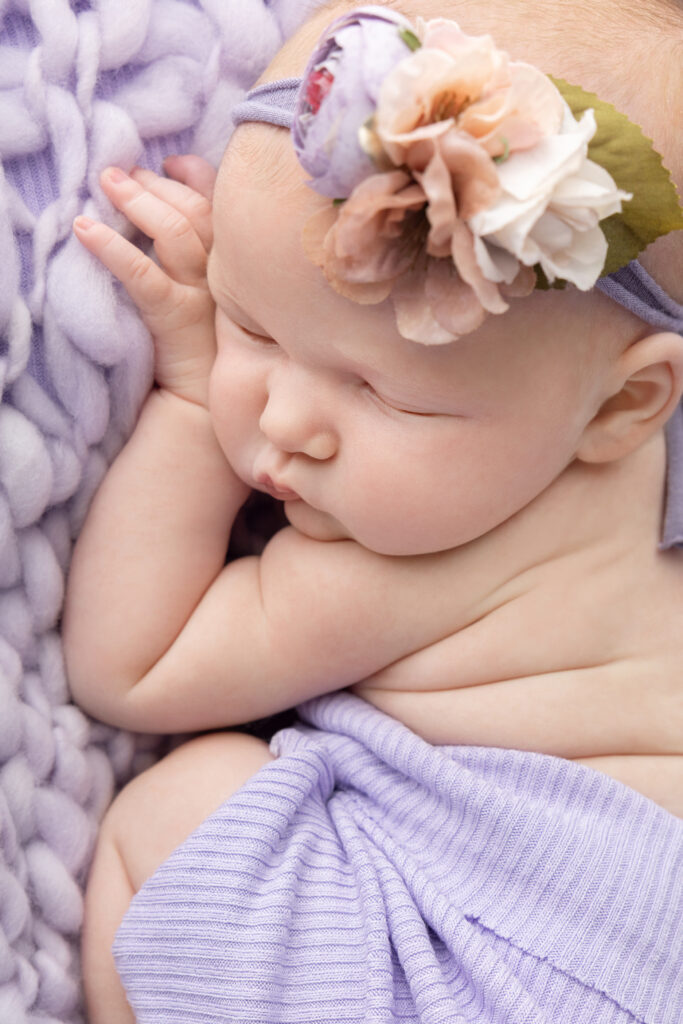A close up image of a newborn baby girl with pale features, wearing a blush, white, and lavender stretchy floral headband. She sleeps on a pale purple plush knit blanket.