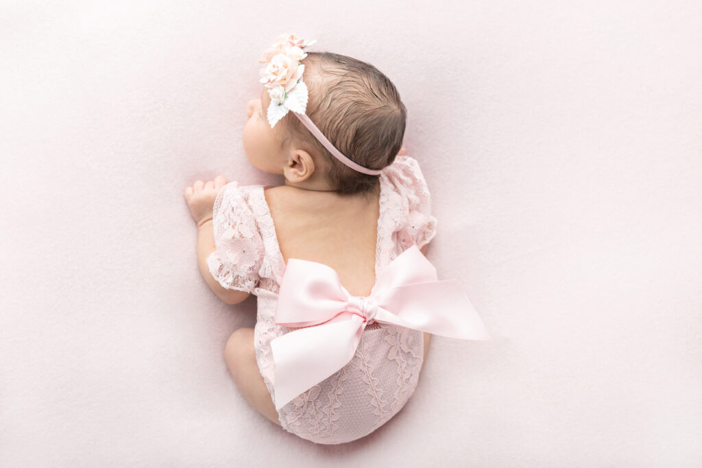 Newborn baby girl Fallon is photographed from above, with her legs tucked up underneath her. Fallon wears a delicate floral headband and her lace outfit is accented by an oversized blush pink bow.