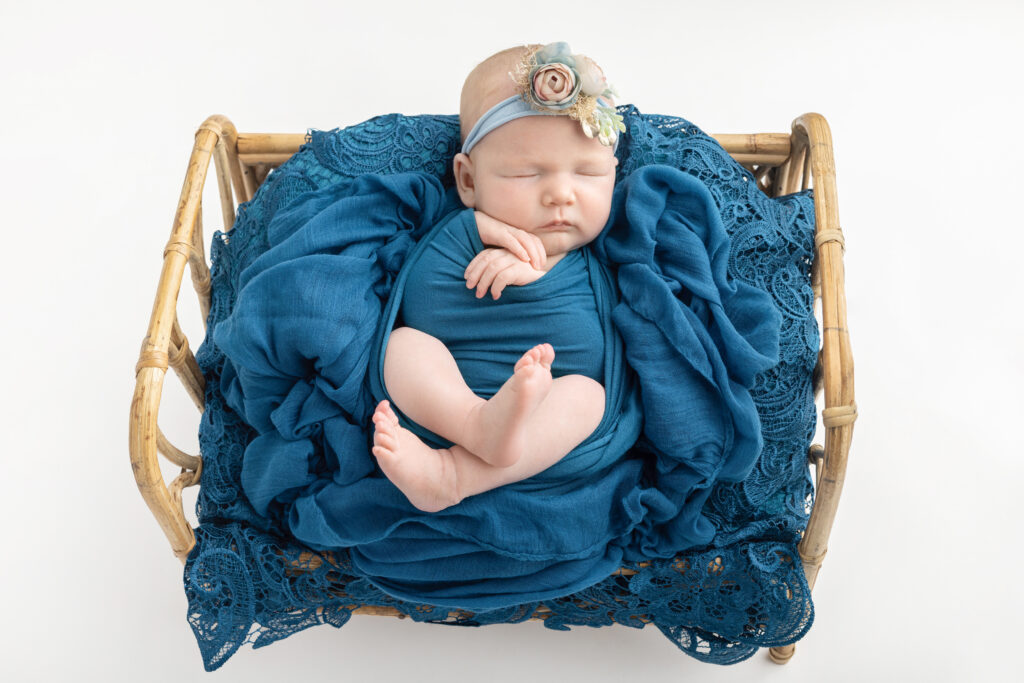 A newborn baby girl sleeps in a rattan basket, loosely wrapped in cerulean and surrounded by a bed of lace in the same color. She wears a pale blue stretchy headband with blush and pale blue flowers.