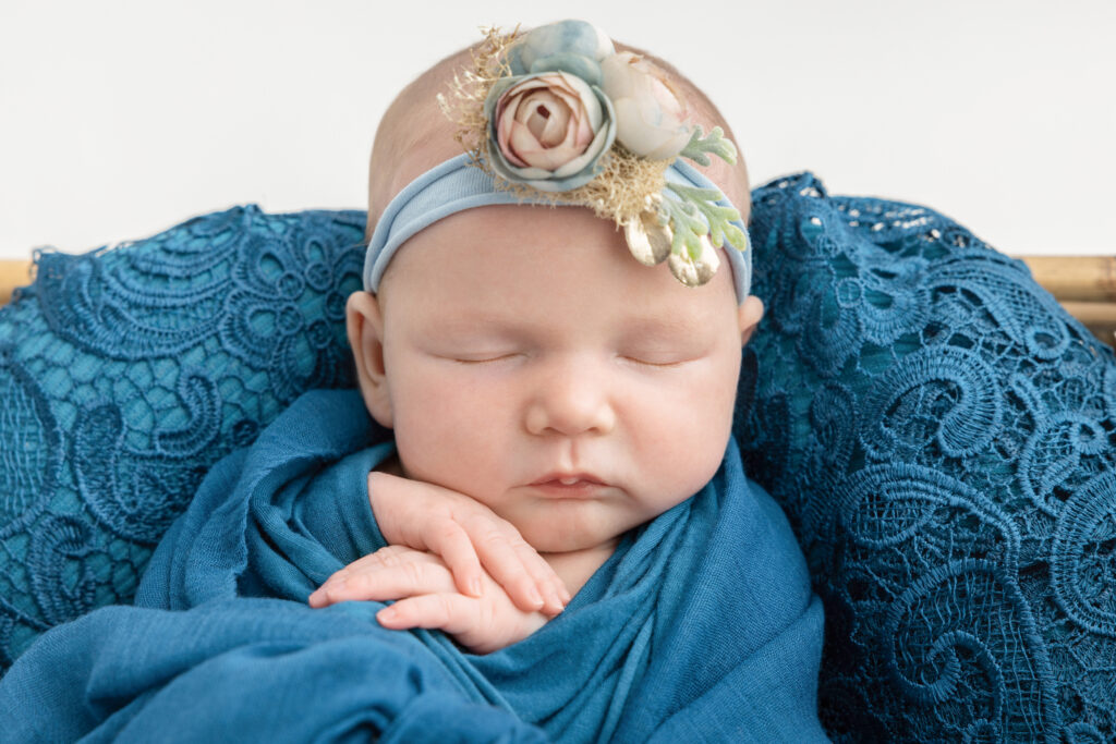 A newborn baby girl sleeps with her hands folded underneath her chin. She is loosely wrapped in cerulean and sleeps on a "bed" of large lace in the same color.