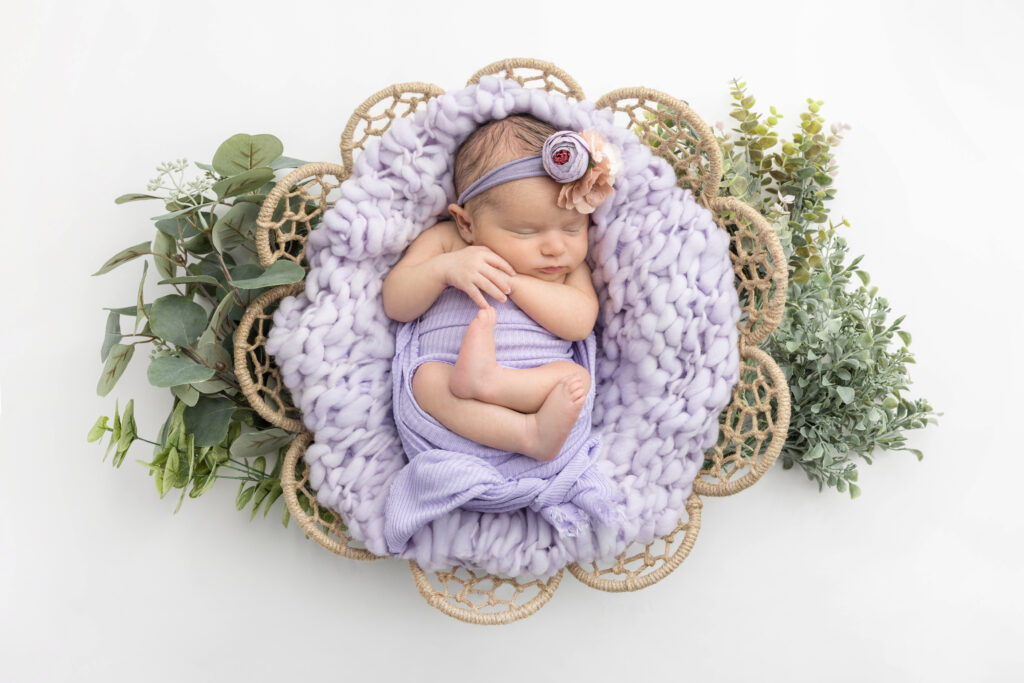 Newborn baby girl Fallon is beautifully swaddled in lilac purple, lying on a chunky, oversized knit blanket in a flower shaped rattan basket, accented by faux eucalyptus and other greenery, against a white backdrop.