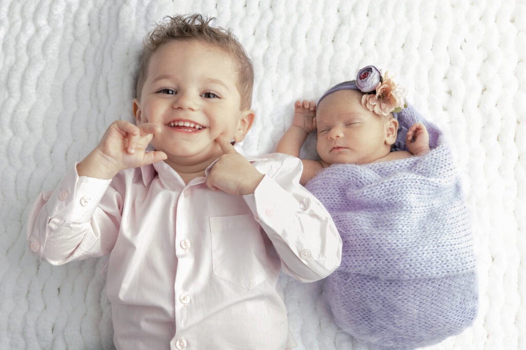 Big brother Cayden, wearing a light pink button-down shirt, shows off his dimples and smiles big as he lies next to his newborn baby sister, Fallon, on a plush white blanket.