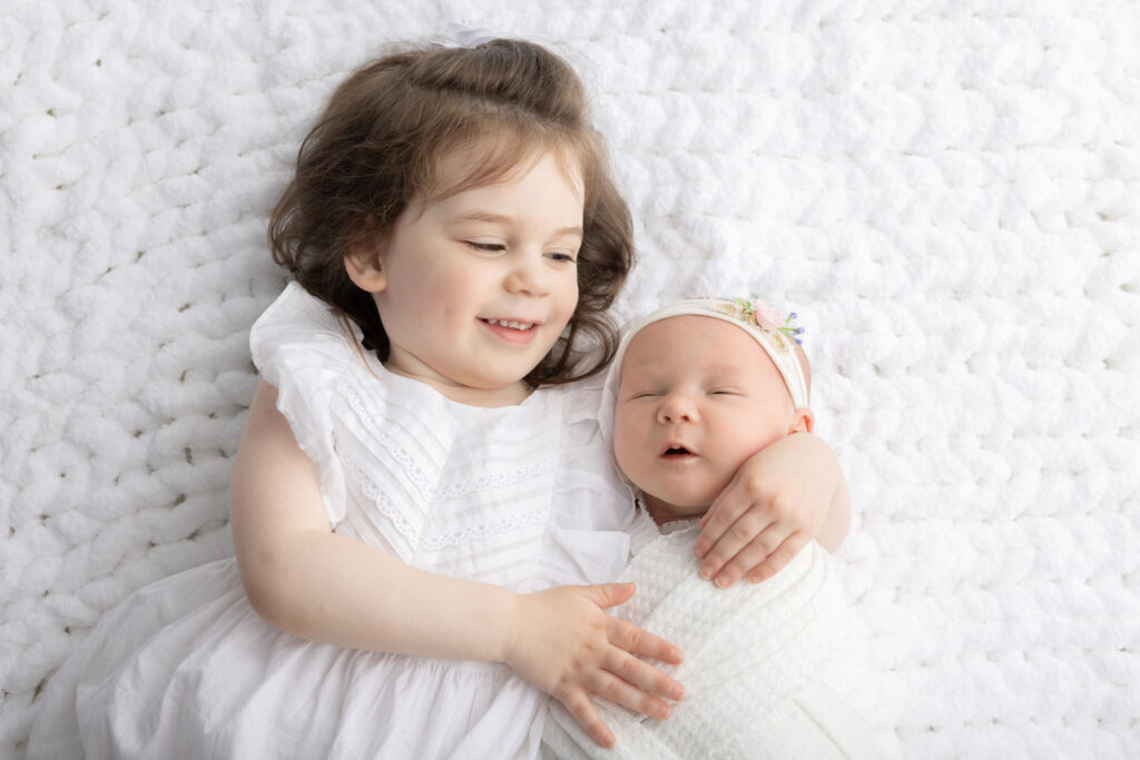 Big sister Blake cradles her baby sister in her arms. The duo are adorned in white, with big sister Blake in a flutter sleeve dress and her baby sister swaddled in white.