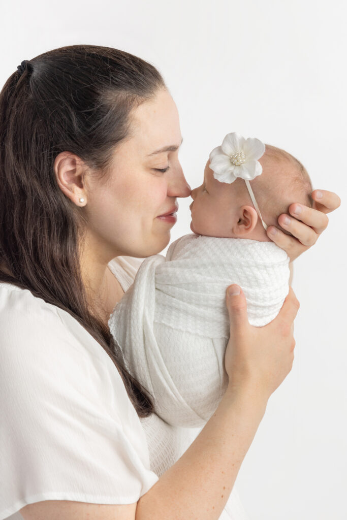 Mama holds her newborn baby girl up to her nose, cradling her in her hands. Mama smiles gently at her darling new baby girl, and they are photographed against a white backdrop. 