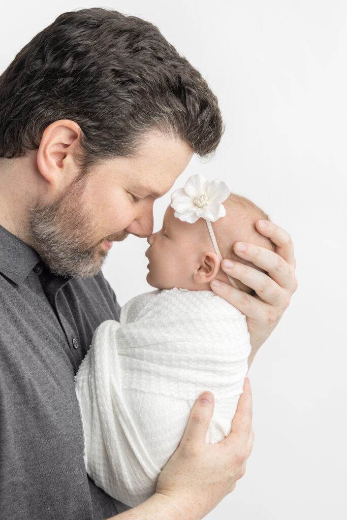 Dad cradles his newborn baby girl in his hands, touching noses with her. Dad has a charcoal polo on, and his daughter is swaddled in white and wears a white floral headband.