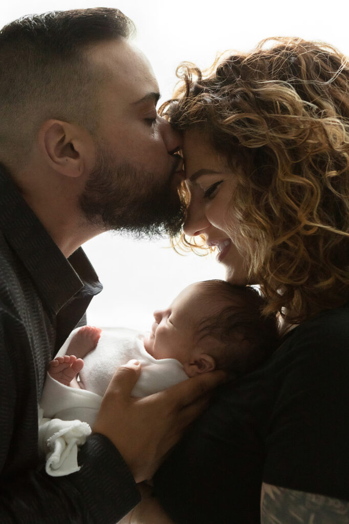 A mother and father hold their newborn baby boy in between them, mother smiling as dad kisses his wife on the forehead. The trio are silhouetted by natural light.