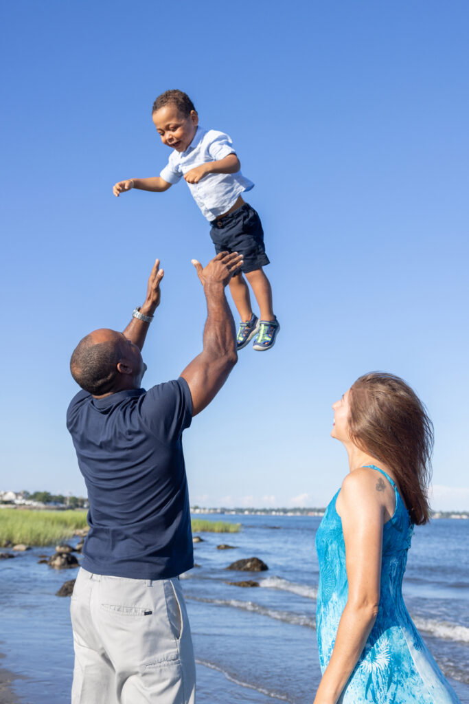 Two year-old Luke's dad tosses him high into the blue sky, arms outstretched to catch him safely. Luke's mom looks up from the side with a smile on her face.