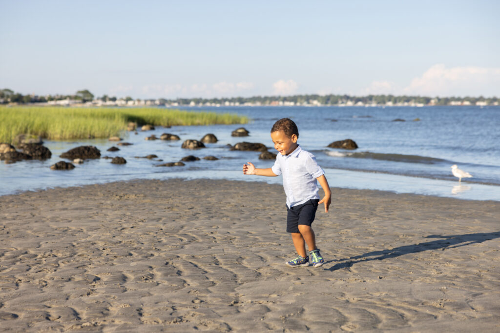 A little boy wearing an oxford shirt with rolled sleeves, navy chino shorts, and navy and green Keen shoes, stomps in the wave patterned sand on the Connecticut shoreline.